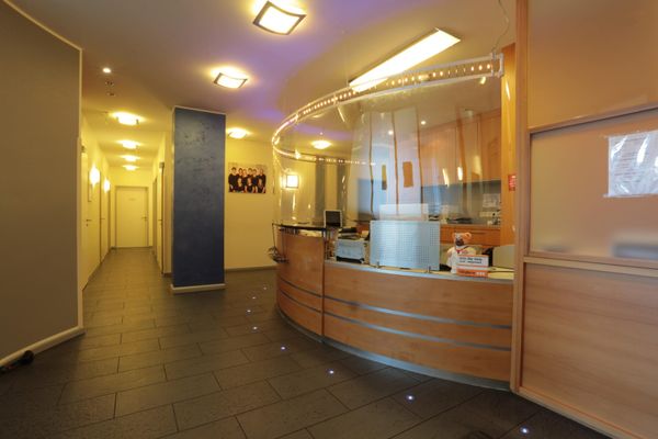 Der helle und moderne Empfangsbereich der Arztpraxis mit einem geschwungenen Holztresen. Ein langer, gut beleuchteter Flur führt von hier zu den Behandlungszimmern.