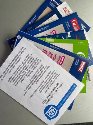 Ein Fächer aus deutschen Zeitschriften (u.a. Bunte, Gala, GEO) und einem Informationsflyer des Hausarzt-Zentrum-Herne. Der Flyer im Vordergrund informiert über die COVID-19-Impfempfehlung für Herbst/Winter 2023 und listet auf, wer sich impfen lassen sollte.
