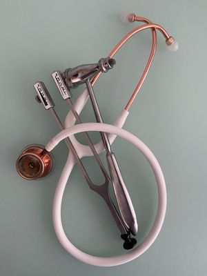 Eine künstlerische Anordnung von medizinischen Instrumenten auf einem mintgrünen Hintergrund. Ein weißes Stethoskop mit kupferfarbenen Metallteilen ist mit einem silbernen neurologischen Reflexhammer und zwei Stimmgabeln verschlungen.